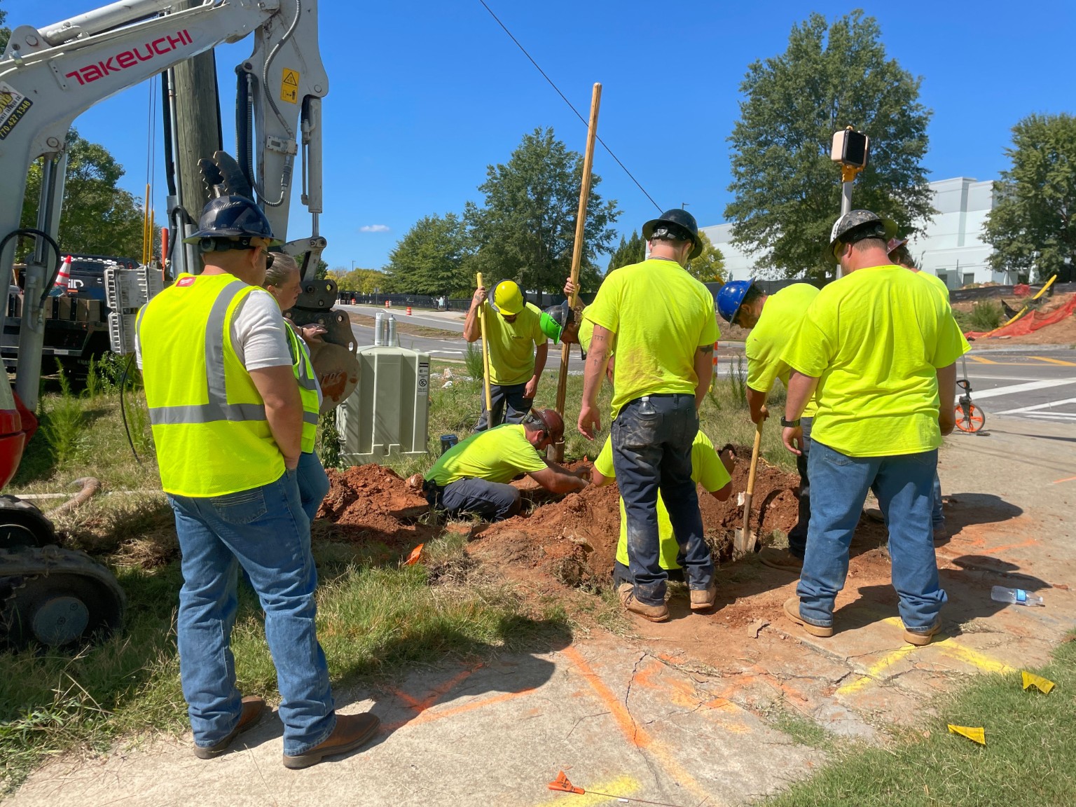 Workers in high-visibility clothing hand-digging beside an excavator, with utility markings on the pavement